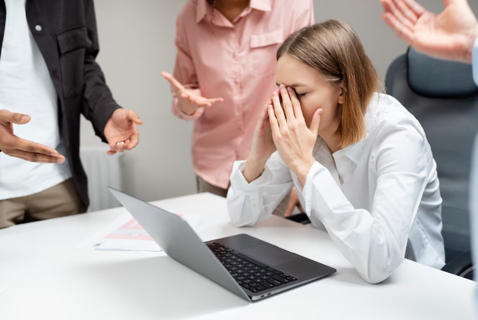 woman sitting near laptop with hands on her face, people standing around her